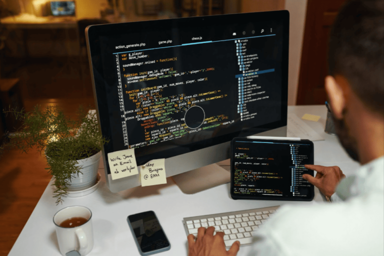 Web developer looking at code on a desktop computer and a tablet next to a cell phone, a cup of tea, and a potted plant.