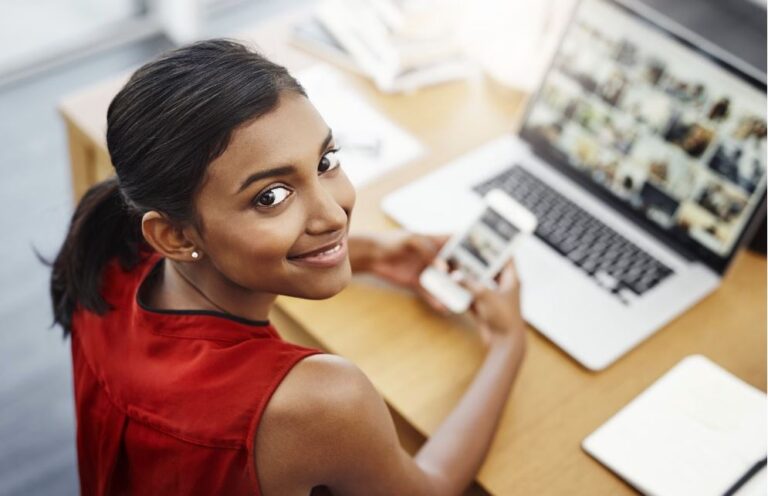 Woman looking at the camera with a cell phone next to a laptop and a wooden desk.