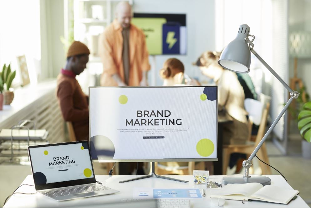 Words “brand marketing” on a desktop and laptop screen with a group of people sitting at a desk in the background.