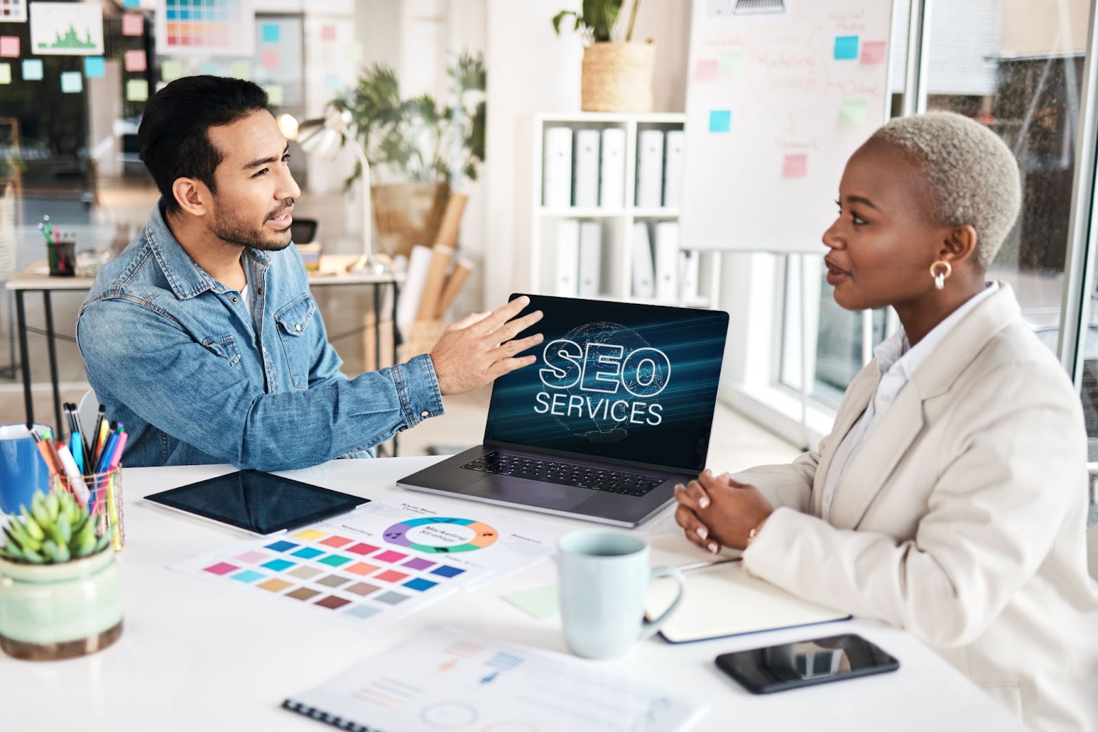 A man and a woman sitting at a desk and looking at a laptop with the words “SEO Services” on the screen.