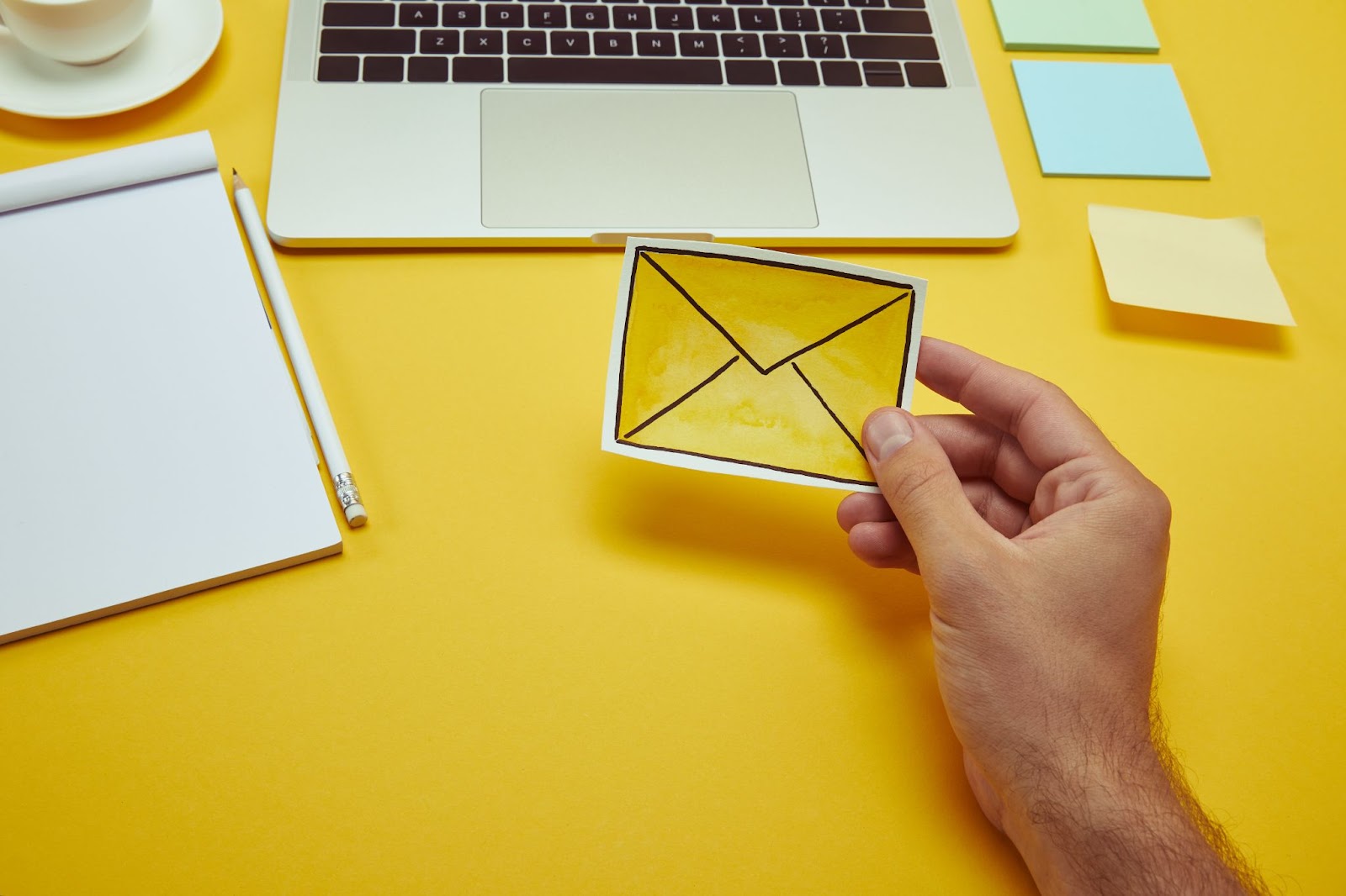 Man holding a drawing of an envelope next to a pad of paper, a laptop, and sticky notes.