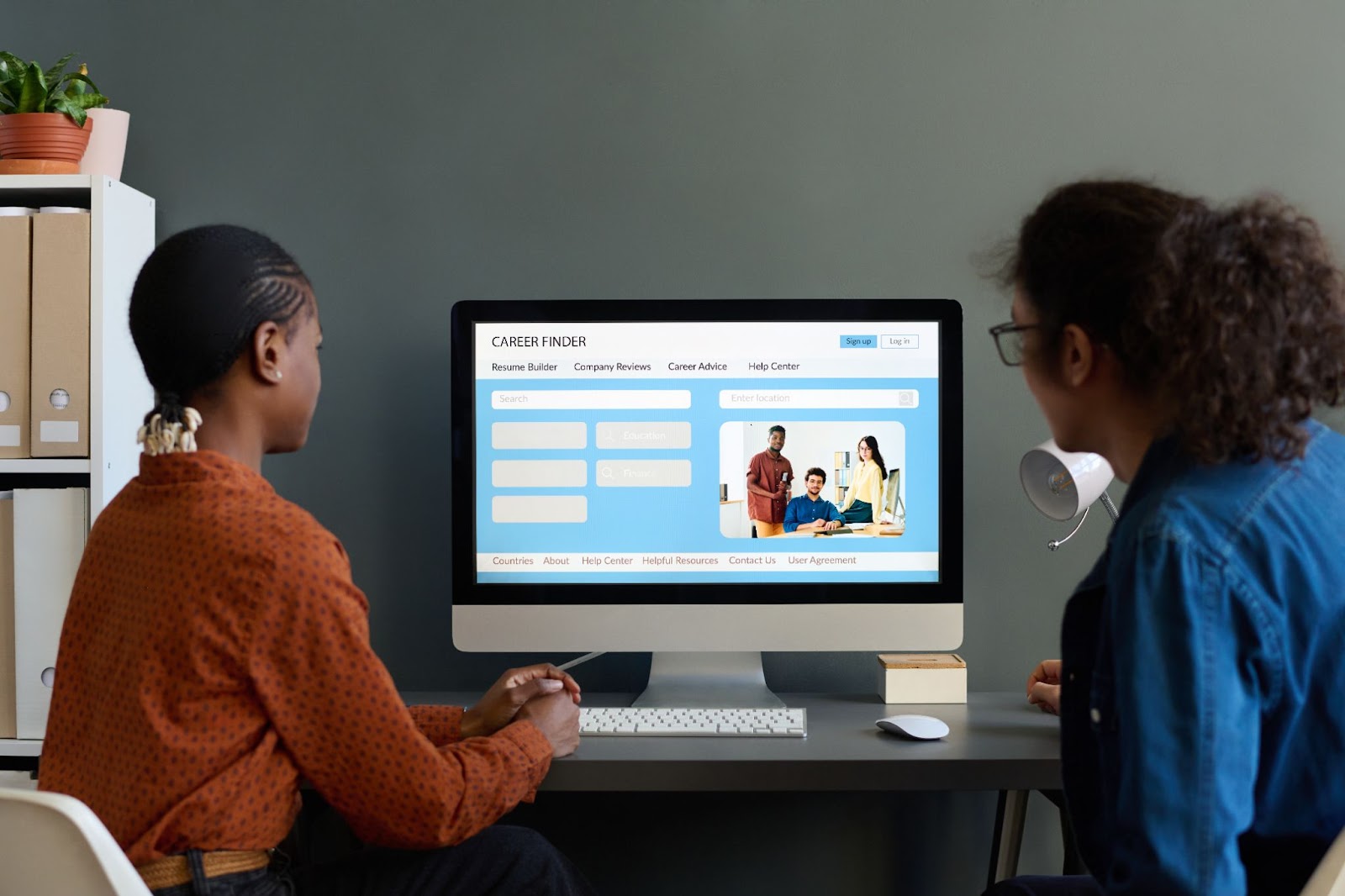 Two people looking at the landing page of a website on a desktop computer on a desk next to a shelf.