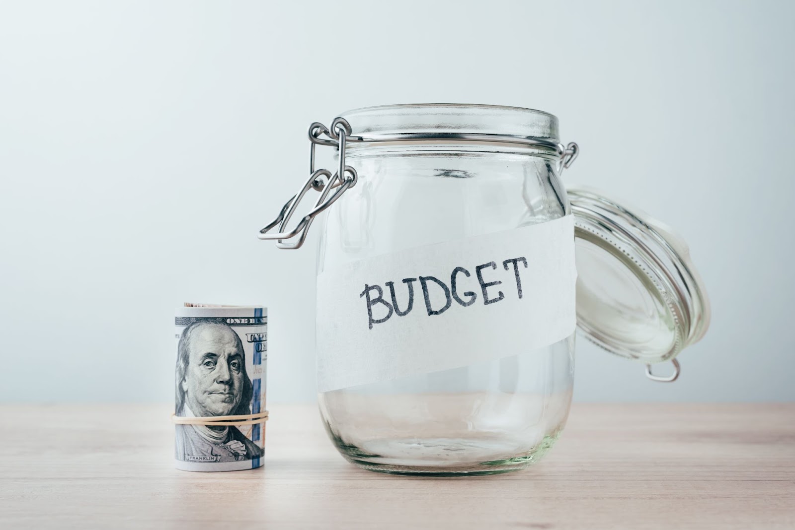 Word “budget” on a glass jar next to a rolled wad of cash on a wooden surface. 