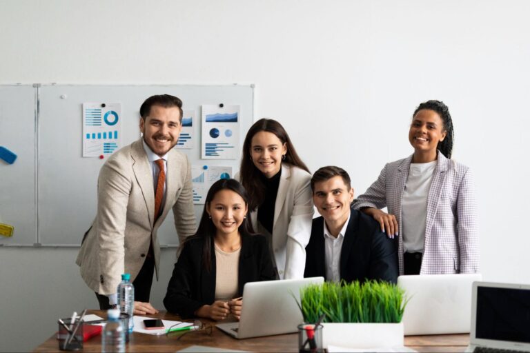 Group of business professionals sitting and standing around a desk with a white board in the background.