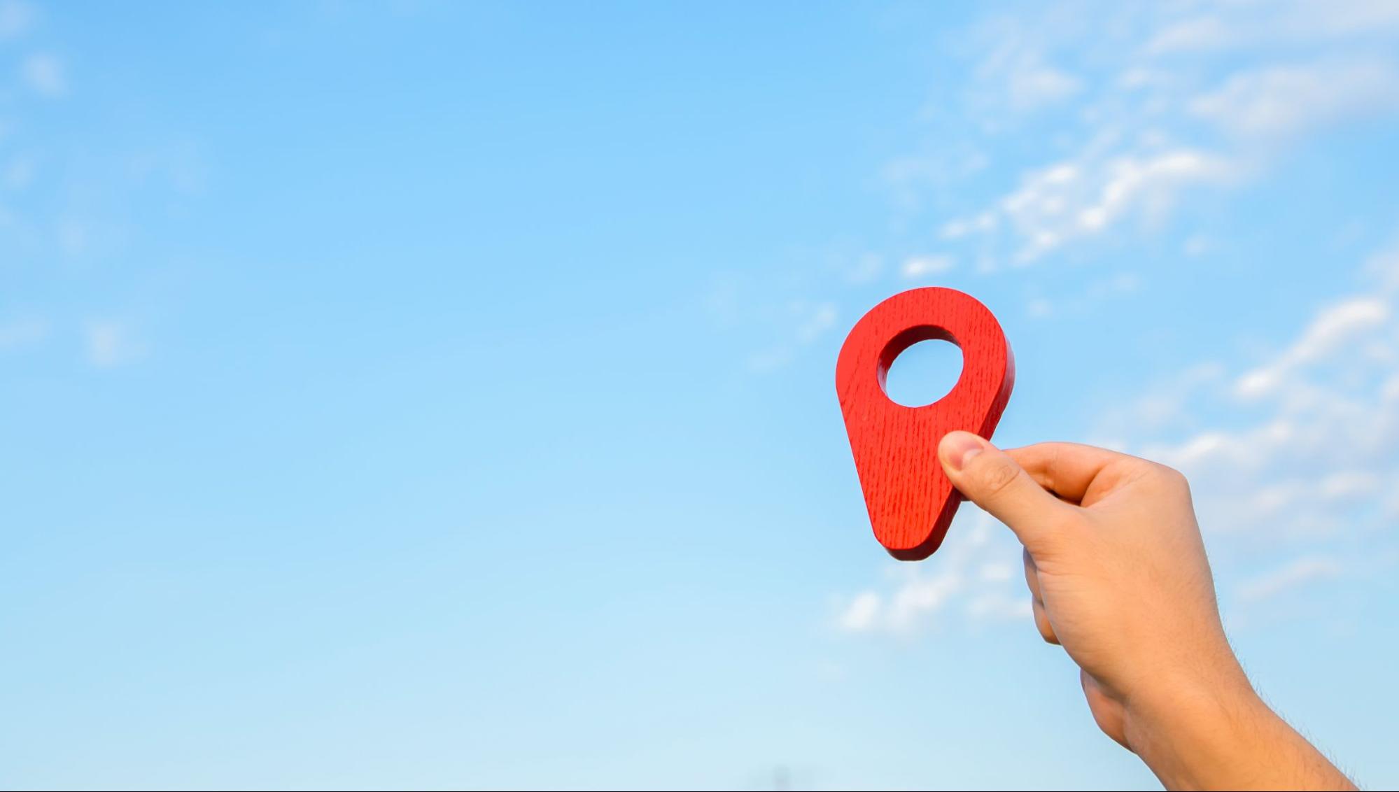 Person holding a wooden location pin logo next to a clear sky with small clouds.