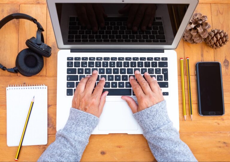 Woman typing on a laptop next to headphones, a pad of paper, pencils, a cell phone, and a pinecone.