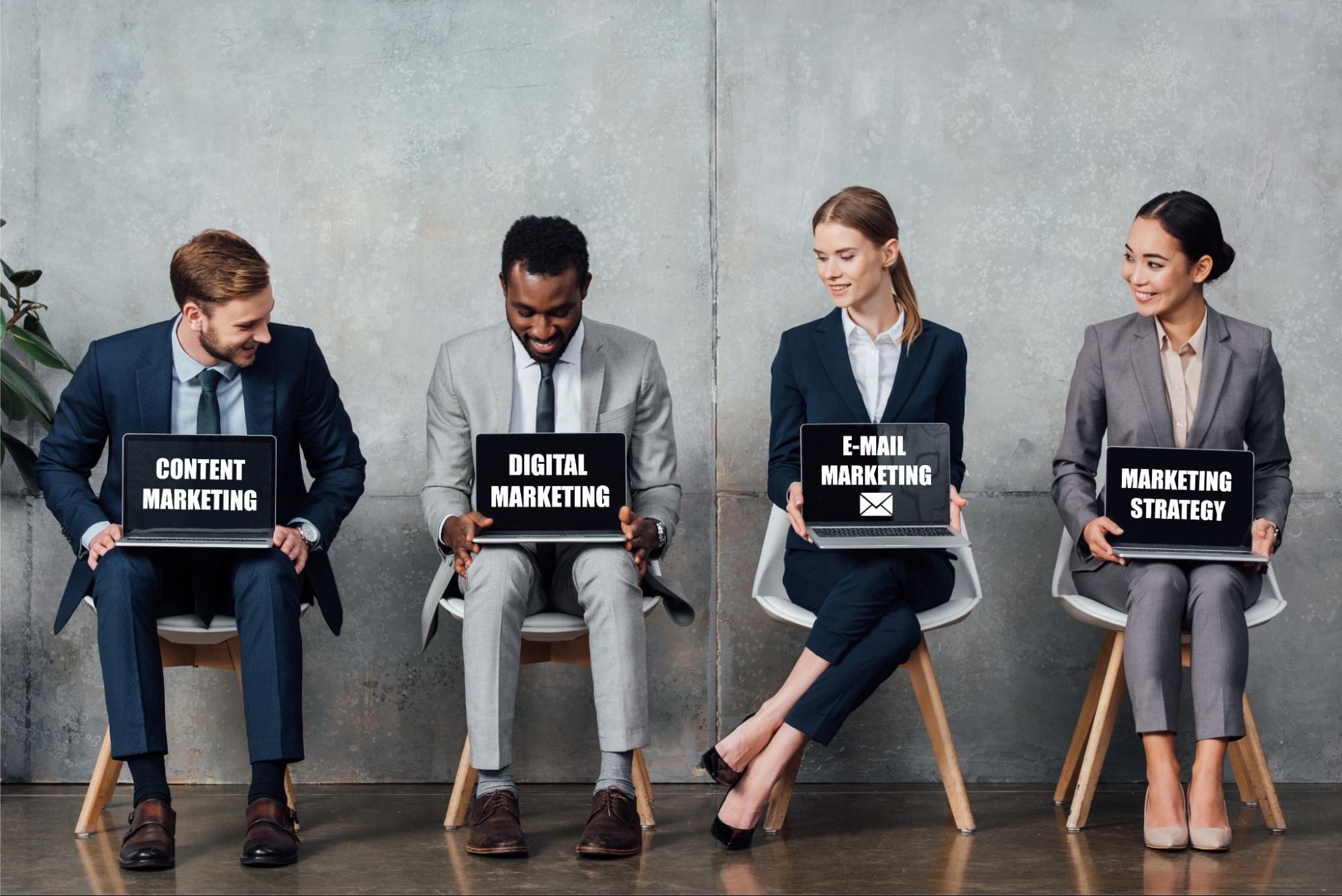 Four business professionals in suits sitting on chairs holding laptops that say different types of marketing strategies.