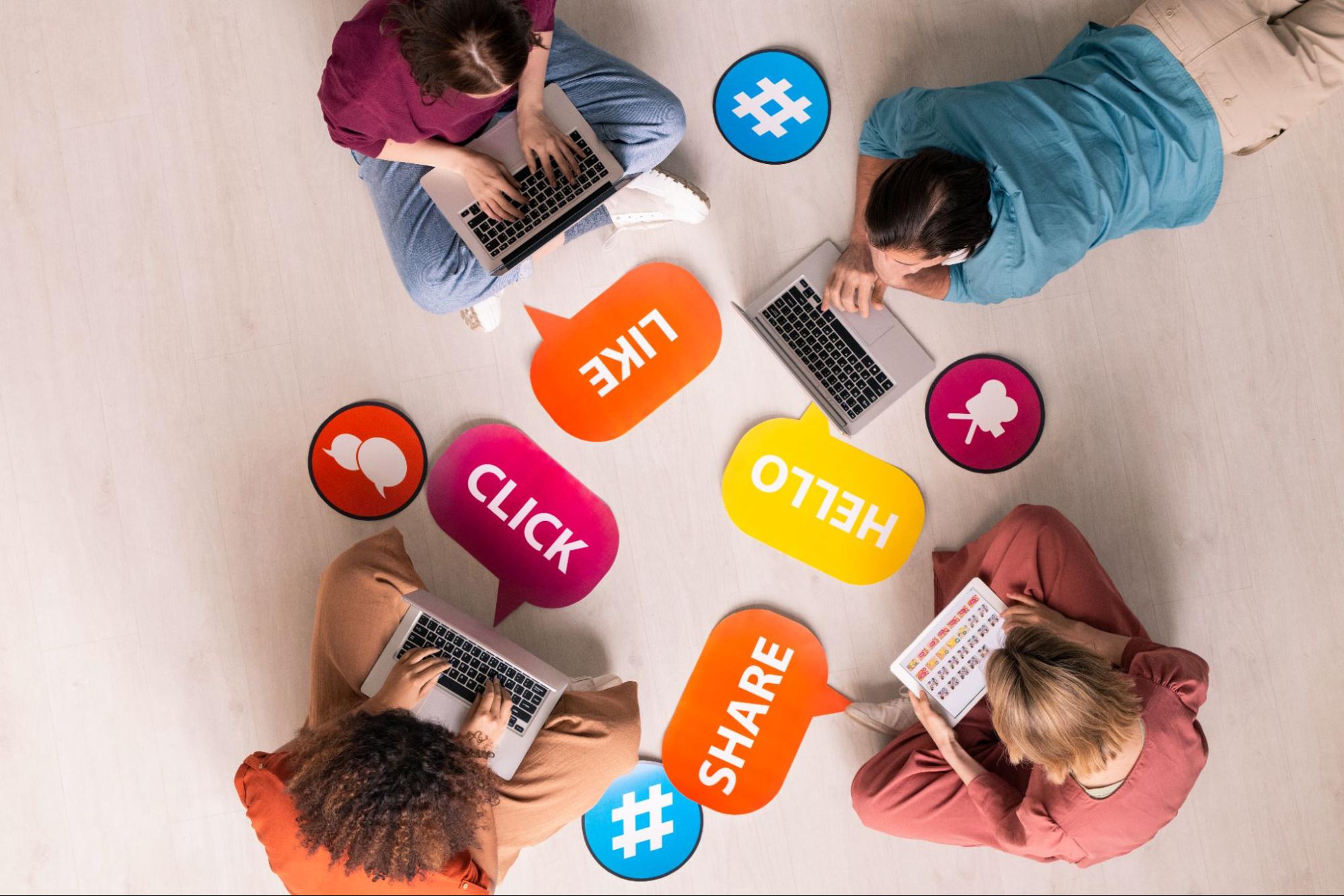 Three women and a man sitting in a circle using laptops next to social media symbols in circles and word bubbles.