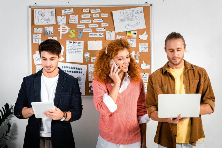 Two men and a woman use technology in front of a corkboard with various pieces of paper and symbols tacked to it.