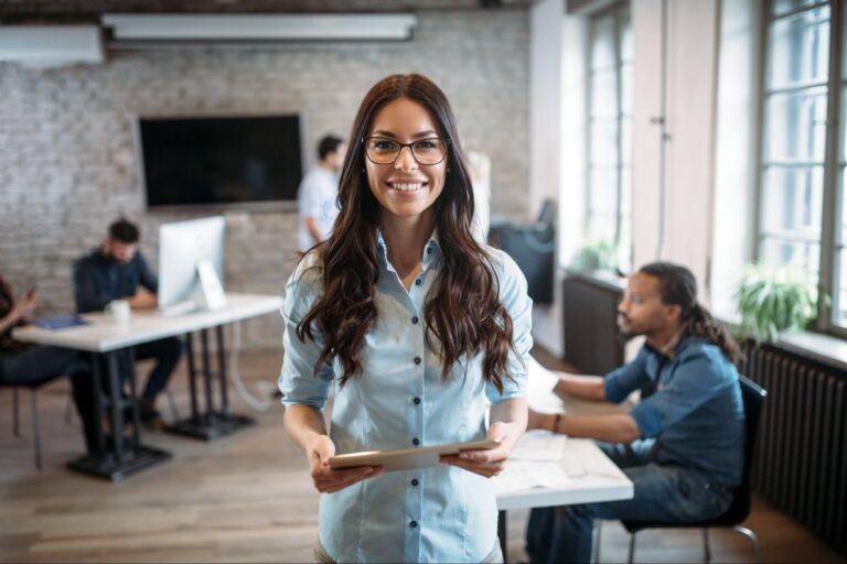 Woman holding an iPad in a modern office with people in the background.