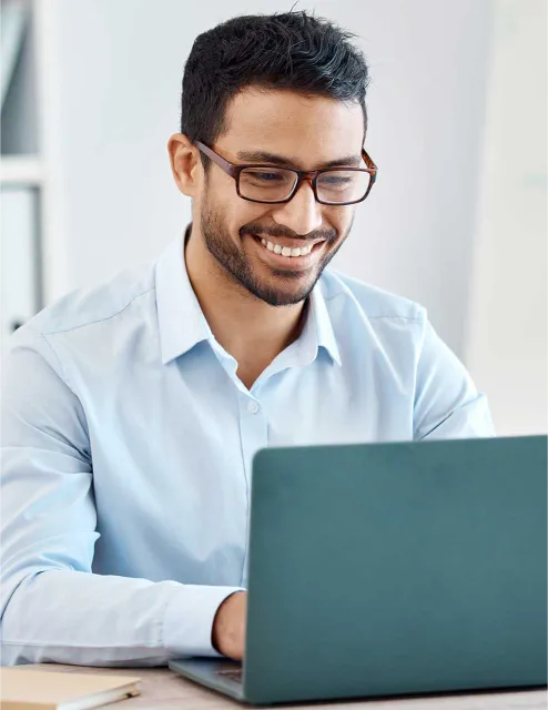 A man wearing glasses and a light blue shirt is smiling while typing on a laptop.